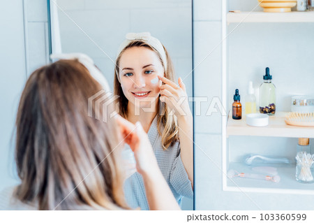 Young woman in bathrobe looking in the mirror, holding bottle with moisturizer cream and applying it on face. Skin hydrating. Cosmetic spa procedures. Beauty self-care at home. Selective focus. Young woman in bathrobe looking in the mirror, holding bottle with moisturizer cream and applying it on face. Skin hydrating. Cosmetic spa procedures. Beauty self-care at home. Selective focus. 103360599