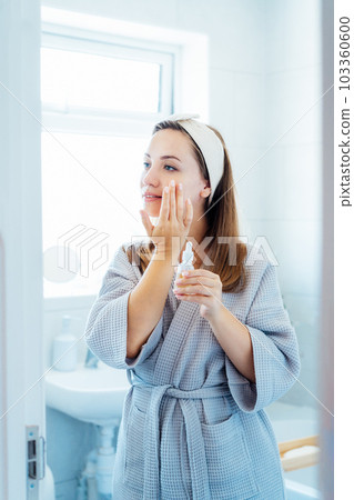Young woman in bathrobe looking in the mirror, holding bottle with hyaluronic acid serum and applying it on face. Skin hydrating. Cosmetic spa procedures. Beauty self-care at home. Selective focus Young woman in bathrobe looking in the mirror, holding bottle with hyaluronic acid serum and applying it on face. Skin hydrating. Cosmetic spa procedures. Beauty self-care at home. Selective focus 103360600