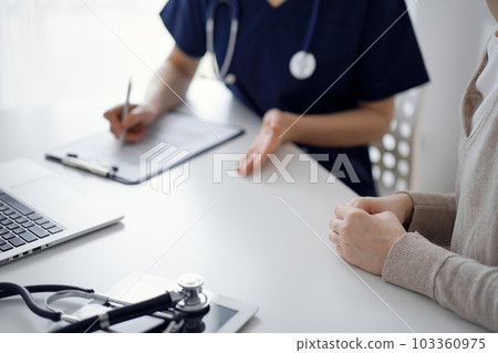 Doctor and patient sitting at the table in clinic while discussing something. The focus is on female patient's hands, close up. Medicine concept Doctor and patient sitting at the table in clinic while discussing something. The focus is on female patient's hands, close up. Medicine concept 103360975