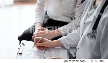 Doctor and patient sitting at sofa in clinic office. The focus is on female physician's hands filling up the medication history record form, close up. Medicine concept Doctor and patient sitting at sofa in clinic office. The focus is on female physician's hands filling up the medication history record form, close up. Medicine concept 103361085