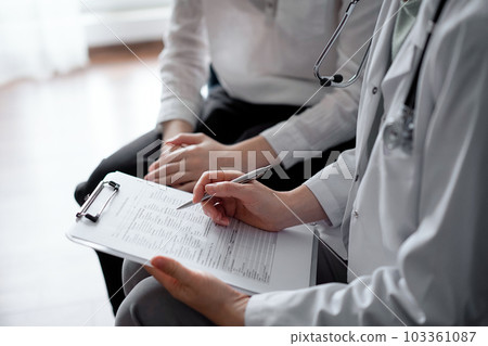 Doctor and patient sitting at sofa in clinic office. The focus is on female physician's hands filling up the medication history record form, close up. Medicine concept Doctor and patient sitting at sofa in clinic office. The focus is on female physician's hands filling up the medication history record form, close up. Medicine concept 103361087