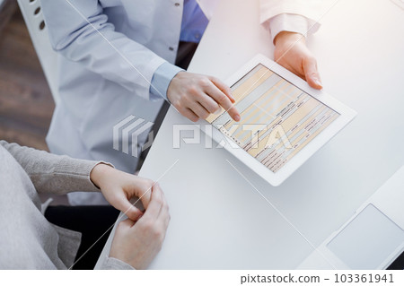 Doctor and patient sitting at the table in clinic. The focus is on female physician's hands using tablet computer, close up. Medicine and healthcare concept Doctor and patient sitting at the table in clinic. The focus is on female physician's hands using tablet computer, close up. Medicine and healthcare concept 103361941