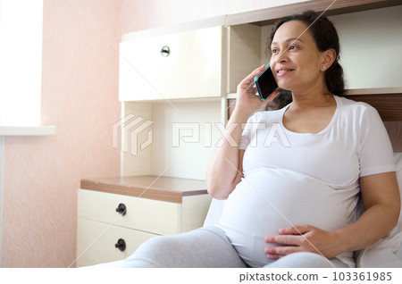 Happy curly haired multi ethnic pregnant woman, smiling looking away, gently caressing her belly while talking on mobile phone, sitting on the bed in cozy light bedroom interior. Pregnancy. Maternity Happy curly haired multi ethnic pregnant woman, smiling looking away, gently caressing her belly while talking on mobile phone, sitting on the bed in cozy light bedroom interior. Pregnancy. Maternity 103361985