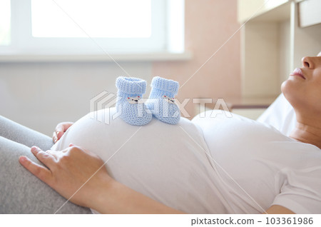 Pregnant woman expecting her future baby, relaxing at home, putting baby booties on her big belly. Pregnancy trimester third, maternity, happy motherhood concept. Close-up advertising shot. Copy space Pregnant woman expecting her future baby, relaxing at home, putting baby booties on her big belly. Pregnancy trimester third, maternity, happy motherhood concept. Close-up advertising shot. Copy space 103361986