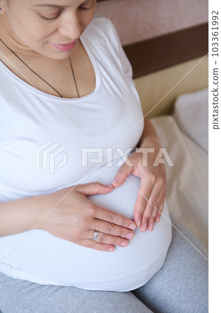 Top view. Charming pregnant woman in white t-shirt, shows a heart made from her fingers over her belly, feeling connection with her future baby, expressing positive emotions of pregnancy and maternity 103361992
