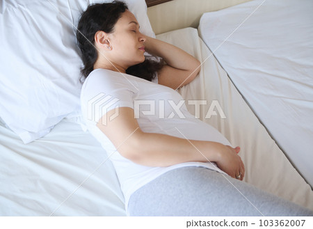 Top view of adult pregnant woman relaxing, sleeping on a bed with her hand on her tummy. Carefree happy pregnancy. Maternity leave. Waiting for the birth of a child. Baby expectation time. Home relax. 103362007