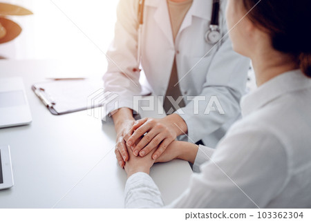 Doctor and patient sitting at the desk in clinic office. The focus is on female physician's hands reassuring woman, close up. Perfect medical service, empathy, and medicine concept 103362304