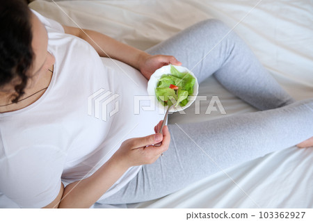 View from above of a pregnant woman, sitting on the bed at home, holding a bowl with fresh salad, enjoying healthy lifestyle during her pregnancy. View from above of a pregnant woman, sitting on the bed at home, holding a bowl with fresh salad, enjoying healthy lifestyle during her pregnancy. 103362927
