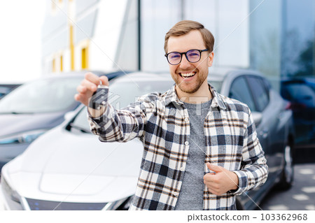 Emotional guy holding key in hand and smiling at camera. Man showing key from his new electric car. Young man buying auto at dealership salon Emotional guy holding key in hand and smiling at camera. Man showing key from his new electric car. Young man buying auto at dealership salon 103362968