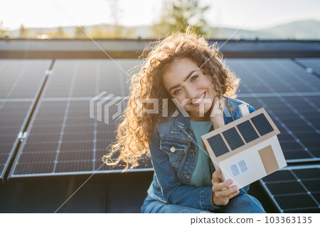 Portrait of young woman on roof with solar panels, holding model of house with photovoltaics. 103363135