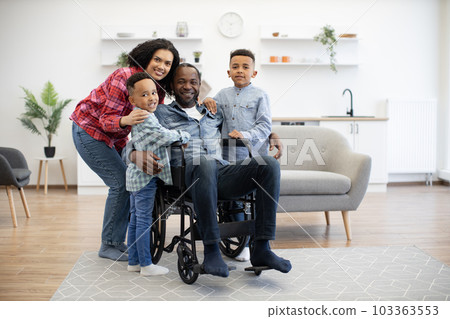 Man in wheelchair hugging wife and kids in living room Man in wheelchair hugging wife and kids in living room 103363553