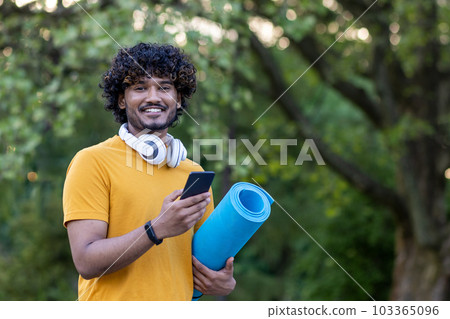 Young hispanic male sports trainer, instructor standing in front of camera in headphones smiling, holding exercise and yoga mat, using phone. 103365096
