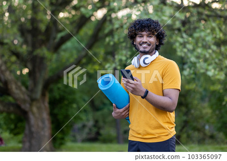 Portrait of hispanic young man standing in park wearing headphones, holding mat and phone. Engaged in physical exercises and sports. Smiling at the camera. 103365097