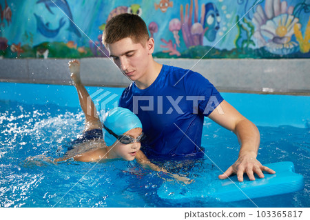 Trainer teaching little boy how to swim in indoor pool with pool floating board 103365817