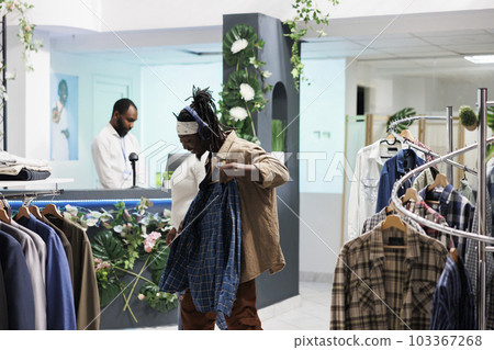 Cheerful buyer checking out shirt while listening to music in headphones in clothing store. African american man in earphones choosing stylish casual apparel in fashion showroom Cheerful buyer checking out shirt while listening to music in headphones in clothing store. African american man in earphones choosing stylish casual apparel in fashion showroom 103367268