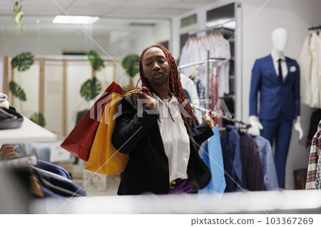 African american woman shopper carrying bags with discounted purchases in shopping mall. Satisfied customer buying apparel for sale and holding paper packages in clothing store African american woman shopper carrying bags with discounted purchases in shopping mall. Satisfied customer buying apparel for sale and holding paper packages in clothing store 103367269