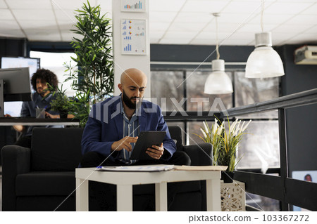 Arab businessman using tablet for digital work in business office. Company man employee scrolling through social media during break while sitting on couch in coworking space 103367272