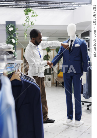 Boutique african american employee checking mannequin wearing stylish jacket and trousers. Man adjusting formal male trendy suit on dummy model in department shopping mall Boutique african american employee checking mannequin wearing stylish jacket and trousers. Man adjusting formal male trendy suit on dummy model in department shopping mall 103367283