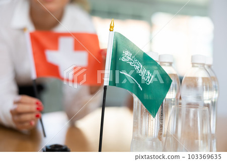Little flag of Saudi Arabia on table and flag of Switzerland put next to it by young woman 103369635