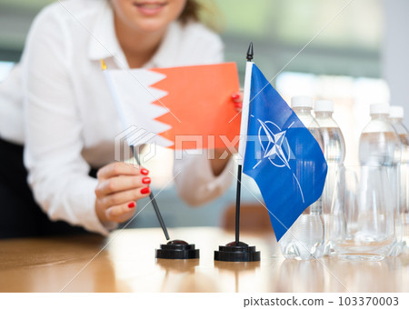 Woman secretary prepares an office for negotiations - she places flags of NATO and Bahrain table 103370003