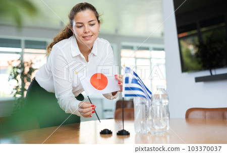 Young lady sets miniatures of flags of Greece and Japan before international negotiations 103370037