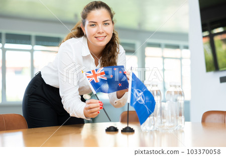 Female secretary places flags of the NATO (OTAN) and Australia on the table before negotiations of top political figures 103370058