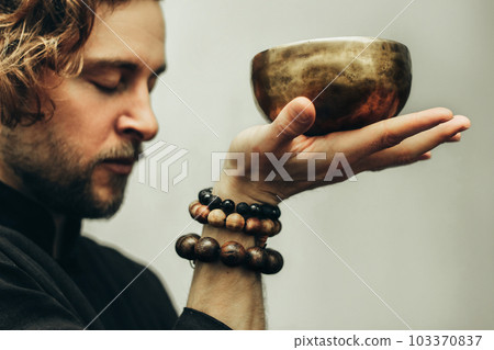 A man with closed eyes listens to the singing bowls. A man holds singing bowls in front of him. The rosary on hand. Music of Tibetan singing bowls. Himalayan bowls. A man with closed eyes listens to the singing bowls. A man holds singing bowls in front of him. The rosary on hand. Music of Tibetan singing bowls. Himalayan bowls. 103370837