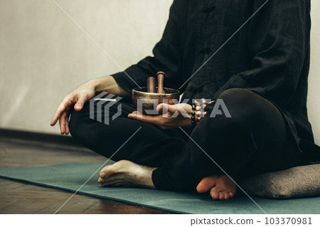 man sits with his legs folded and holding a copper singing bowl with sticks in his hands. Relaxation and meditation. Alternative medicine tibetan singing bowls man sits with his legs folded and holding a copper singing bowl with sticks in his hands. Relaxation and meditation. Alternative medicine tibetan singing bowls 103370981