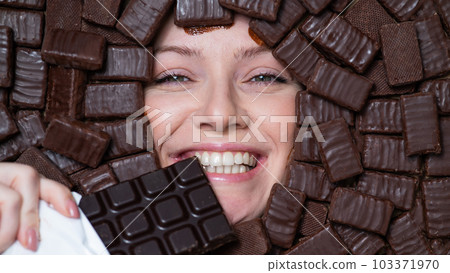 The face of a caucasian woman surrounded by chocolates. The girl eats a bar of chocolate. 103371970