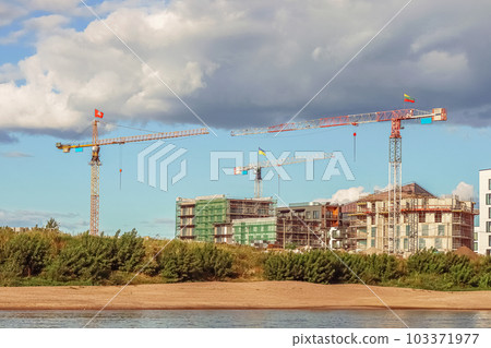 Construction site with three cranes with three flags Ukrainian, Lithuanian and Lithuanian coat of arms building residential buildings on river bank with sandy beach 103371977