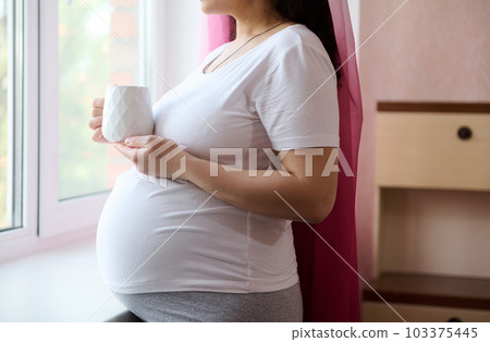 Close-up of a happy pregnant woman in white t-shirt, standing by window at home bedroom, looking out the window, relaxing with a cup of herbal drink. Pregnancy trimester third. Happy maternity leave Close-up of a happy pregnant woman in white t-shirt, standing by window at home bedroom, looking out the window, relaxing with a cup of herbal drink. Pregnancy trimester third. Happy maternity leave 103375445