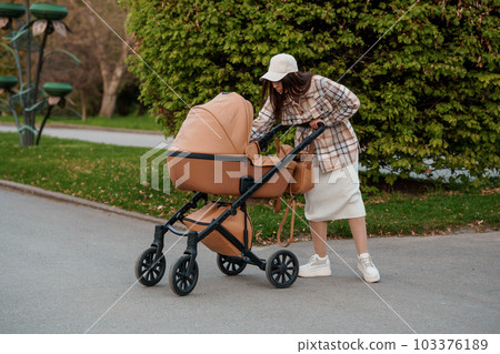 Portrait of a beautiful girl. Close-up photo of a beautiful girl in a plaid shirt and a white cap. The girl stands near the brown baby carriage. Mother walking with a small child 103376189