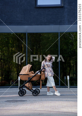 Portrait of a beautiful girl. Close-up photo of a beautiful girl in a plaid shirt and a white cap. The girl stands near the brown baby carriage. Mother walking with a small child Portrait of a beautiful girl. Close-up photo of a beautiful girl in a plaid shirt and a white cap. The girl stands near the brown baby carriage. Mother walking with a small child 103376203