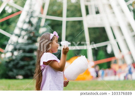 Child girl in an amusement park in the summer eats cotton candy and smiles happily. The concept of summer holidays and school holidays 103376889