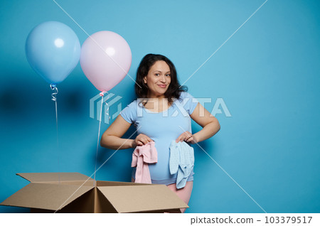 Happy pregnant woman with big belly, holding newborn clothes, smiling at camera, posing with balloons of pink and blue pastel color, isolated blue background. Baby shower. Gender reveal party concept Happy pregnant woman with big belly, holding newborn clothes, smiling at camera, posing with balloons of pink and blue pastel color, isolated blue background. Baby shower. Gender reveal party concept 103379517