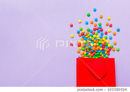 shopping paper gift bag in corner full of assorted traditional candies falling out on colored background with copy space. Happy Holidays sale concept shopping paper gift bag in corner full of assorted traditional candies falling out on colored background with copy space. Happy Holidays sale concept 103380504