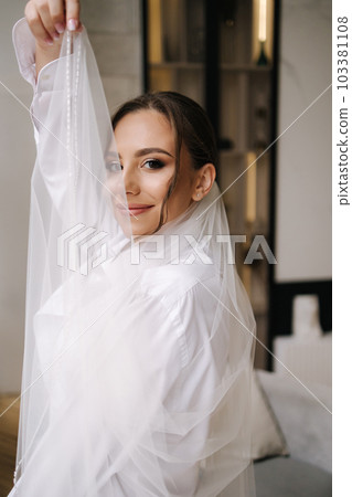 Beautiful bride with long bridal veil dressed in grooms shirt stand in front of big window at home Beautiful bride with long bridal veil dressed in grooms shirt stand in front of big window at home 103381108