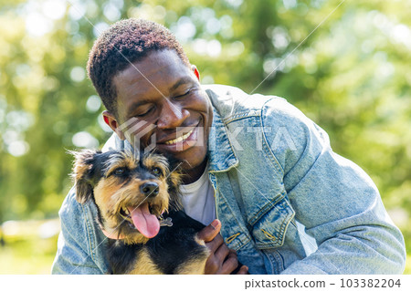 latin american man walking with his cute dog at sunny day in city park lawn on the grass 103382204