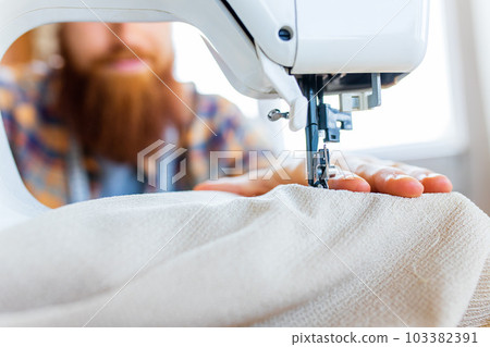 handsome redhaired man with long beard sews at a sewing machine at home studio handsome redhaired man with long beard sews at a sewing machine at home studio 103382391