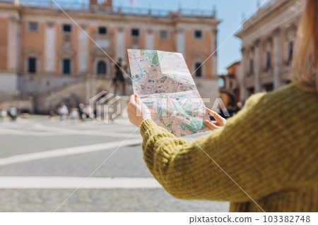 Blonde Woman with paper map is walking on a sunny day. Piazza del Campidoglio on Capitoline Hill, Rome, Italy. Concept of travel, tourism and vacation in city. Blonde Woman with paper map is walking on a sunny day. Piazza del Campidoglio on Capitoline Hill, Rome, Italy. Concept of travel, tourism and vacation in city. 103382748