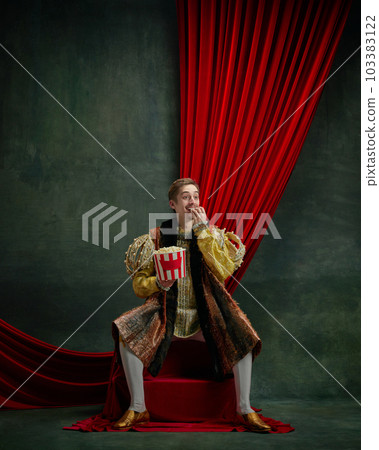 Portrait of smiling young man, royal person, prince eating popcorn and watching comedy movie against dark green, vintage background 103383122