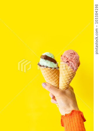 A cone of refreshing ice cream in a waffle cup against a yellow wall. The concept of vacation, vacation at sea. A woman holds ice cream in her hands. Vertical photo. A cone of refreshing ice cream in a waffle cup against a yellow wall. The concept of vacation, vacation at sea. A woman holds ice cream in her hands. Vertical photo. 103384198