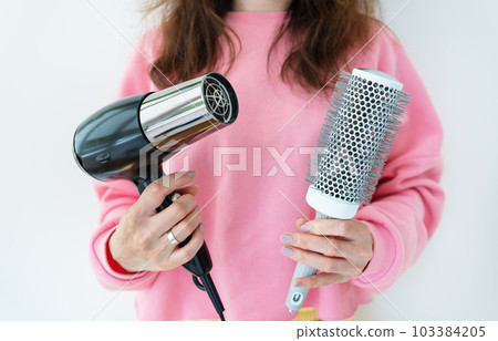 The girl holds a hair dryer and a brush in her hands. Drying long brown hair with a round brush for styling unruly hair. Beauty saloon. 103384205