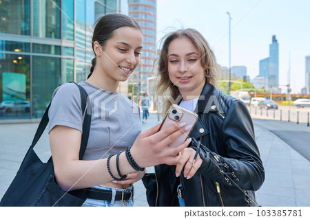 Two smiling female students looking at smartphone, urban modern background 103385781