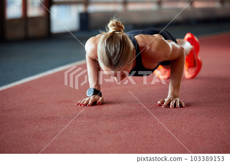 Young athlete girl is warming up before the race at the stadium. A slender blonde in a black tracksuit is doing push-ups on the track. Sports and recreation. 103389153