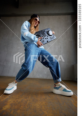 A young pretty long-haired DJ girl in a blue sweater, jeans and a black baseball cap poses with a black DJ mixing console and imitates energetic dance moves. Studio shot, gray background. 103389165