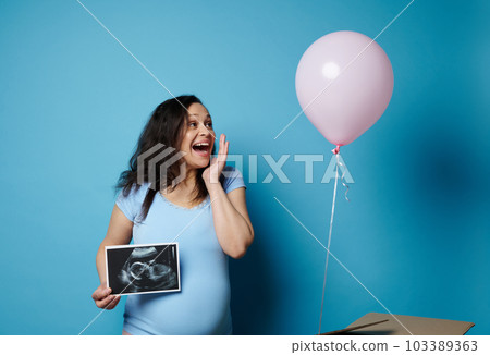 Pregnant woman experiencing joy while watching a pink balloon flying out of a cardboard box, smiling, holding ultrasound scan image of her future child, isolated on blue backdrop. Gender reveal party Pregnant woman experiencing joy while watching a pink balloon flying out of a cardboard box, smiling, holding ultrasound scan image of her future child, isolated on blue backdrop. Gender reveal party 103389363