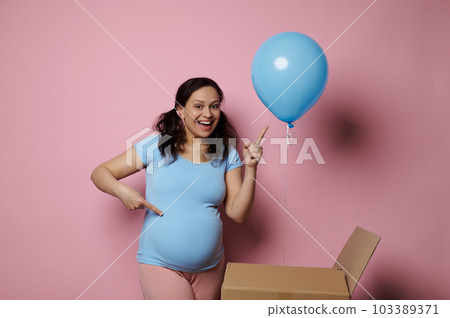 Expectant future mom, pregnant woman smiling and pointing finger at blue balloon, she's expecting a baby boy, isolated on pink background. Gender reveal party concept. Pregnancy. Maternity. Motherhood 103389371
