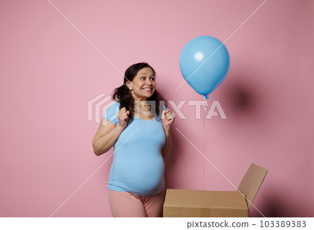Happy pregnant woman, experiencing joy and happiness discovering a blue baloon at gender reveal party. Gravid mother expecting a baby boy, standing isolated on pink background, dressed in blue t-shirt Happy pregnant woman, experiencing joy and happiness discovering a blue baloon at gender reveal party. Gravid mother expecting a baby boy, standing isolated on pink background, dressed in blue t-shirt 103389383