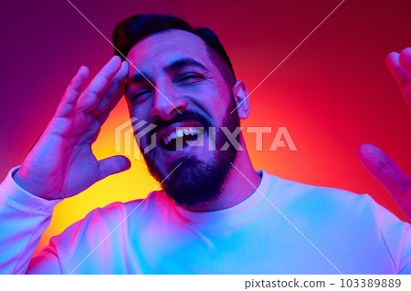 Portrait of bearded handsome man cheerfully smiling against gradient multicolored studio background in neon light. Happiness, positive vibe Portrait of bearded handsome man cheerfully smiling against gradient multicolored studio background in neon light. Happiness, positive vibe 103389889